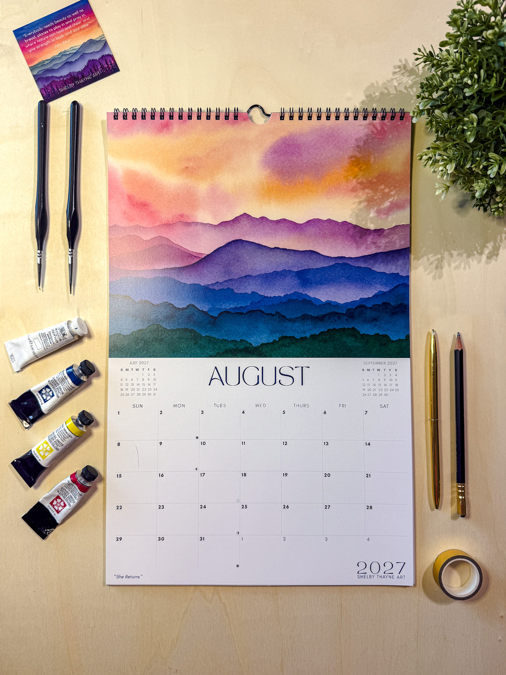 A spiral-bound August 2027 wall calendar featuring a colorful watercolor mountain landscape, styled in a flat lay on a light wood surface with paint tubes, brushes, gold pens, tape, and a small plant.
