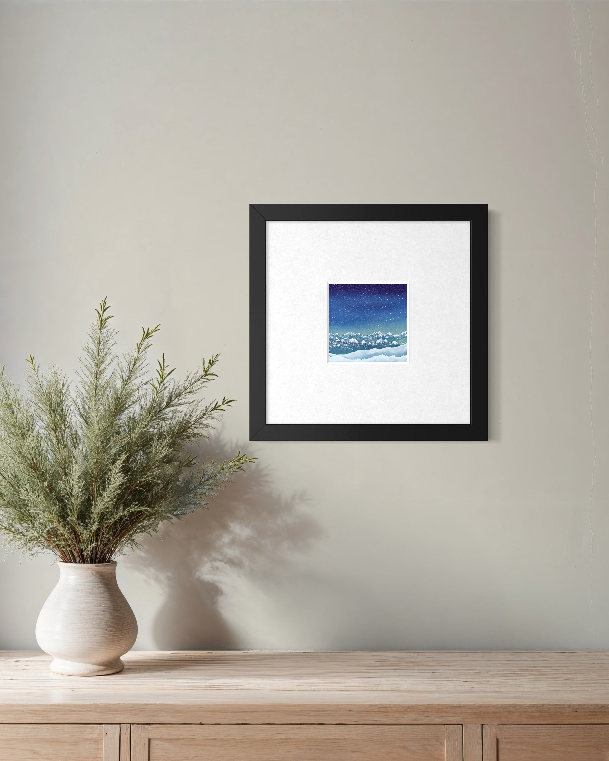 Framed artwork on a wall above a wooden surface with a vase of plants.