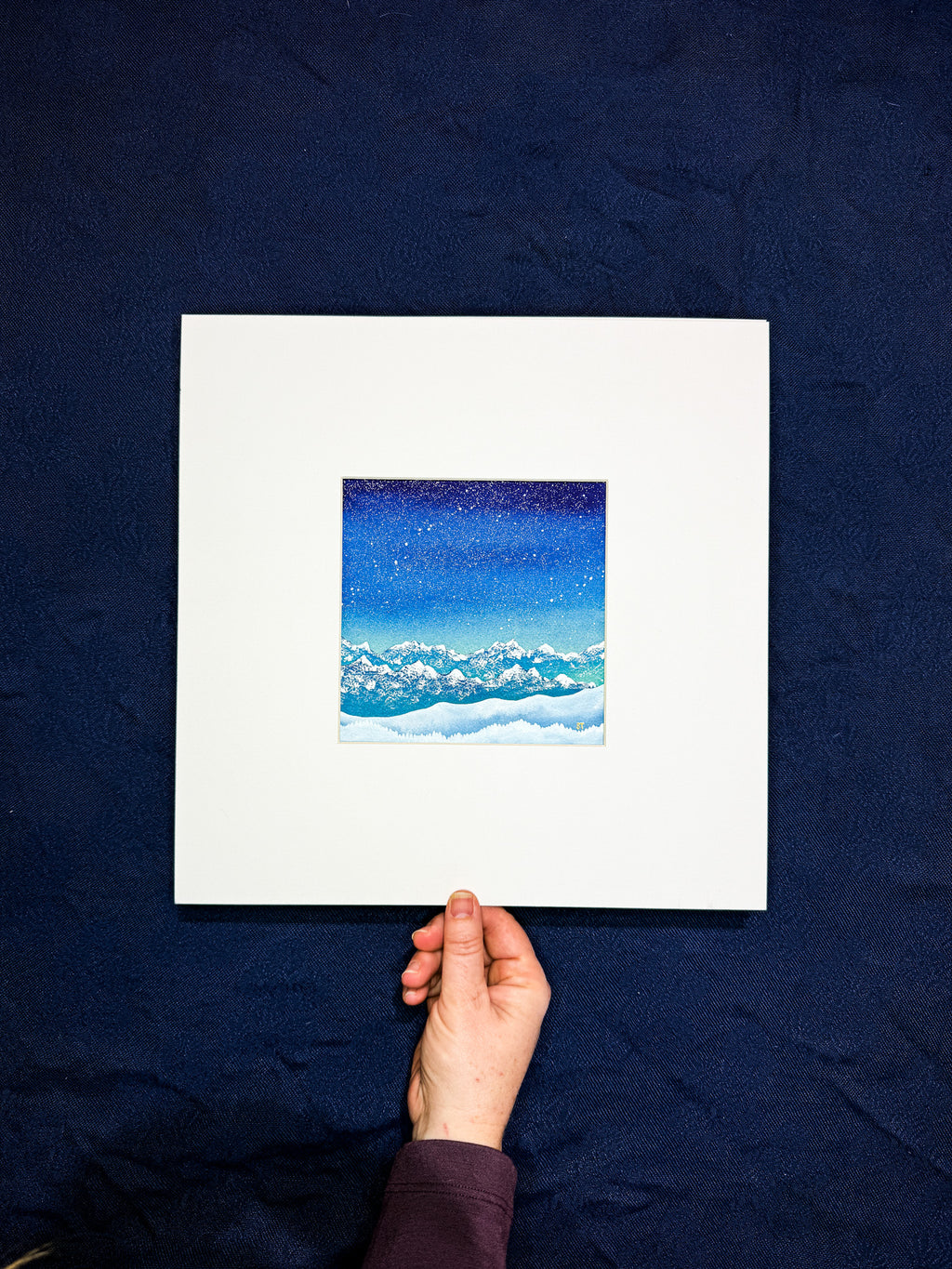 Hand holding a framed artwork of a night sky with clouds against a dark blue background