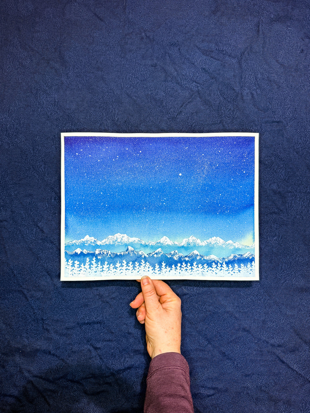 Hand holding a piece of paper with a starry night sky and landscape drawing against a dark blue fabric background.