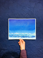 Hand holding a piece of paper with a starry night sky and landscape drawing against a dark blue fabric background.