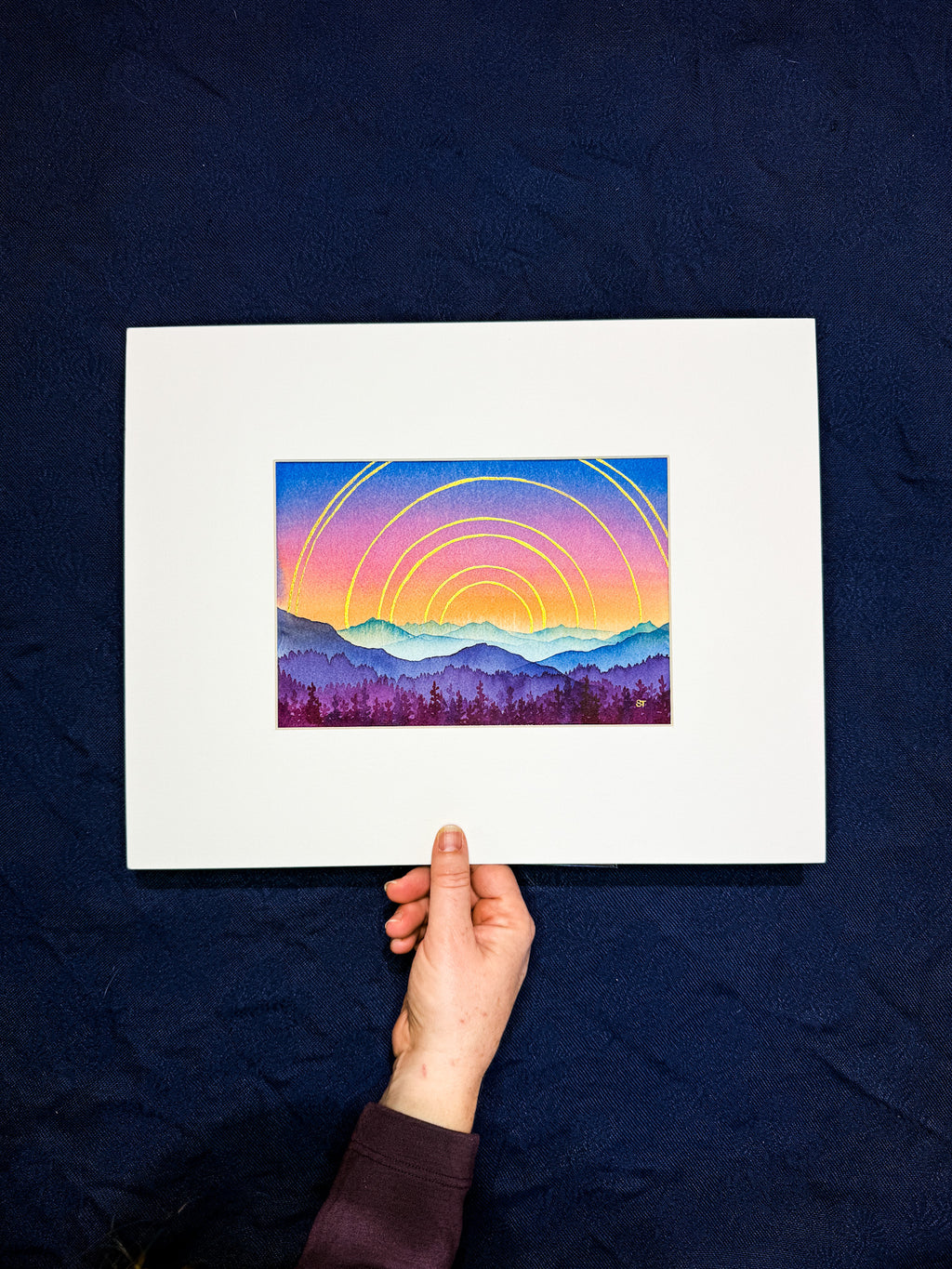 Hand holding a framed artwork of a colorful sunset over mountains on a dark blue background