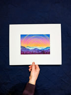 Hand holding a framed artwork of a colorful sunset over mountains on a dark blue background