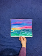 Hand holding a small watercolor painting of a colorful landscape against a blue textured background