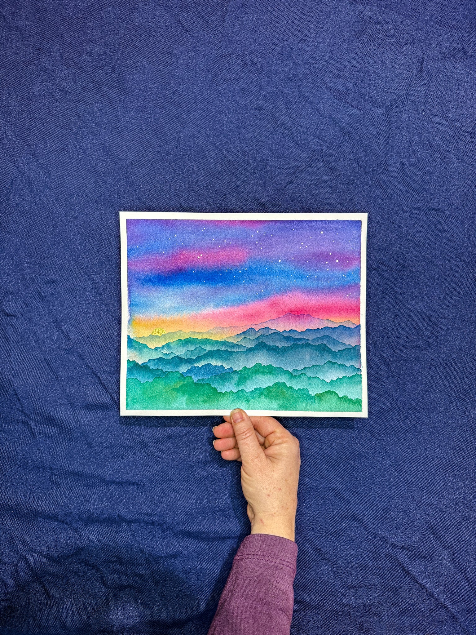 Hand holding a small watercolor painting of a colorful landscape against a blue textured background