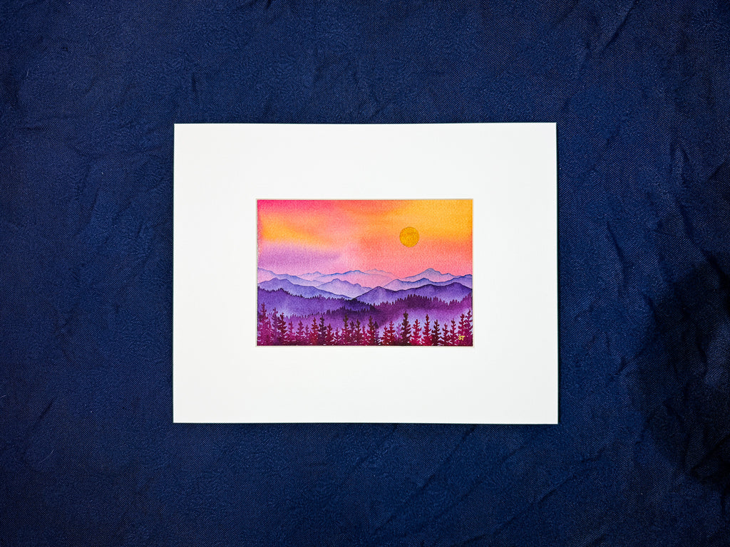 Framed artwork of a sunset over mountains and trees on a textured blue background