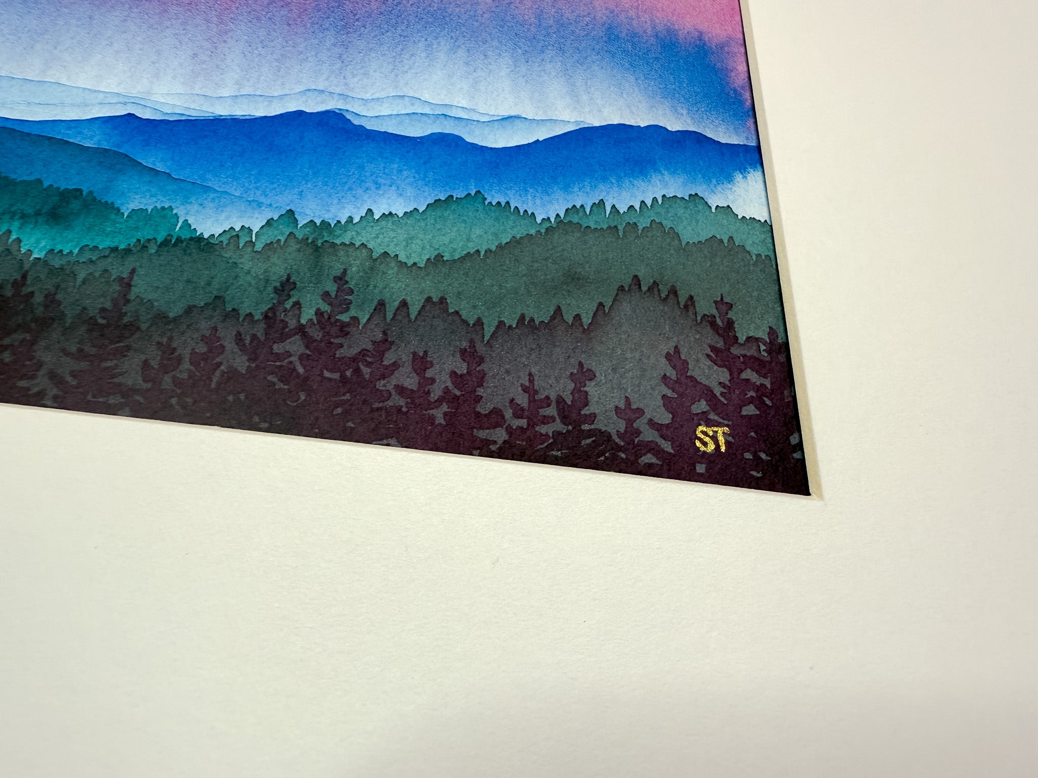 Artistic depiction of a forest with watercolor-style mountains and artist initials in gold framed in a white mat 