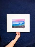 Hand holding a matted watercolor painting of mountains against a dark blue background