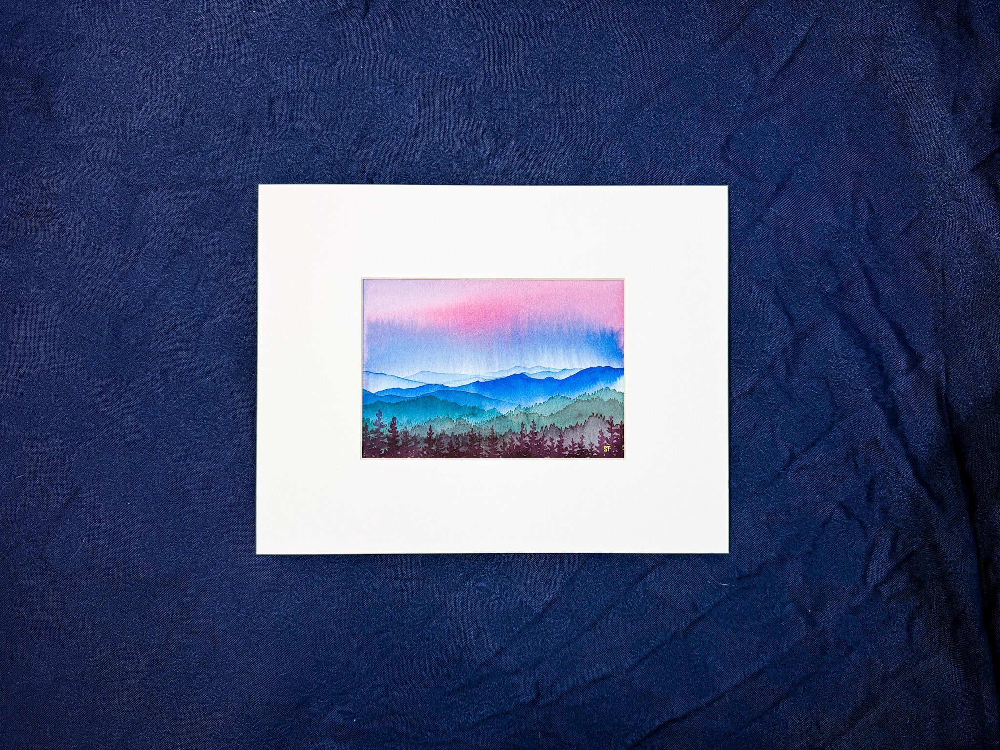 Small painting of a mountain landscape watercolor painting in a white mat on a textured blue fabric background