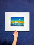 Hand holding a framed artwork of a mountain landscape with trees against a blue background