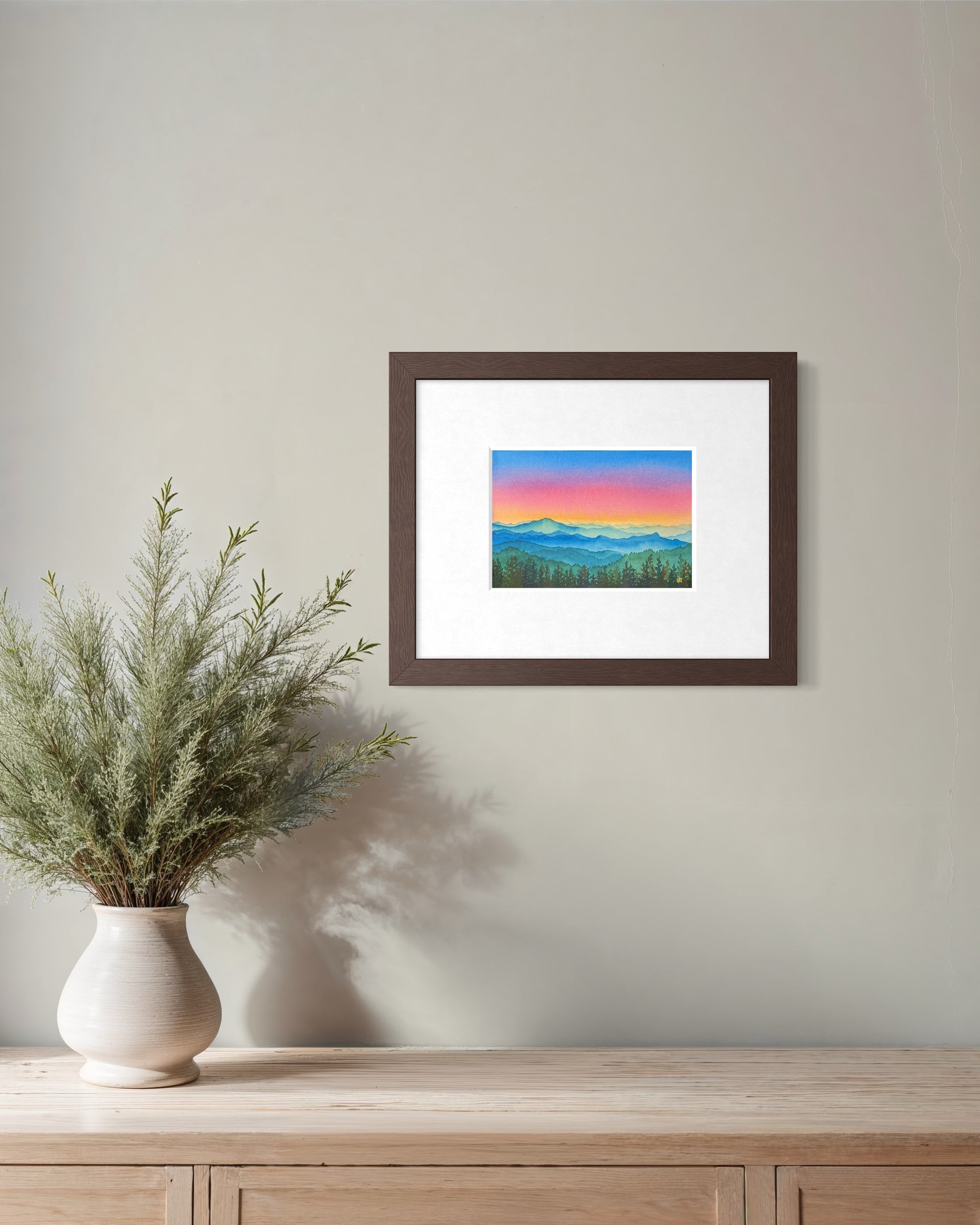 Framed artwork of a mountain landscape with a colorful sky on a wall above a wooden cabinet with a vase of plants.