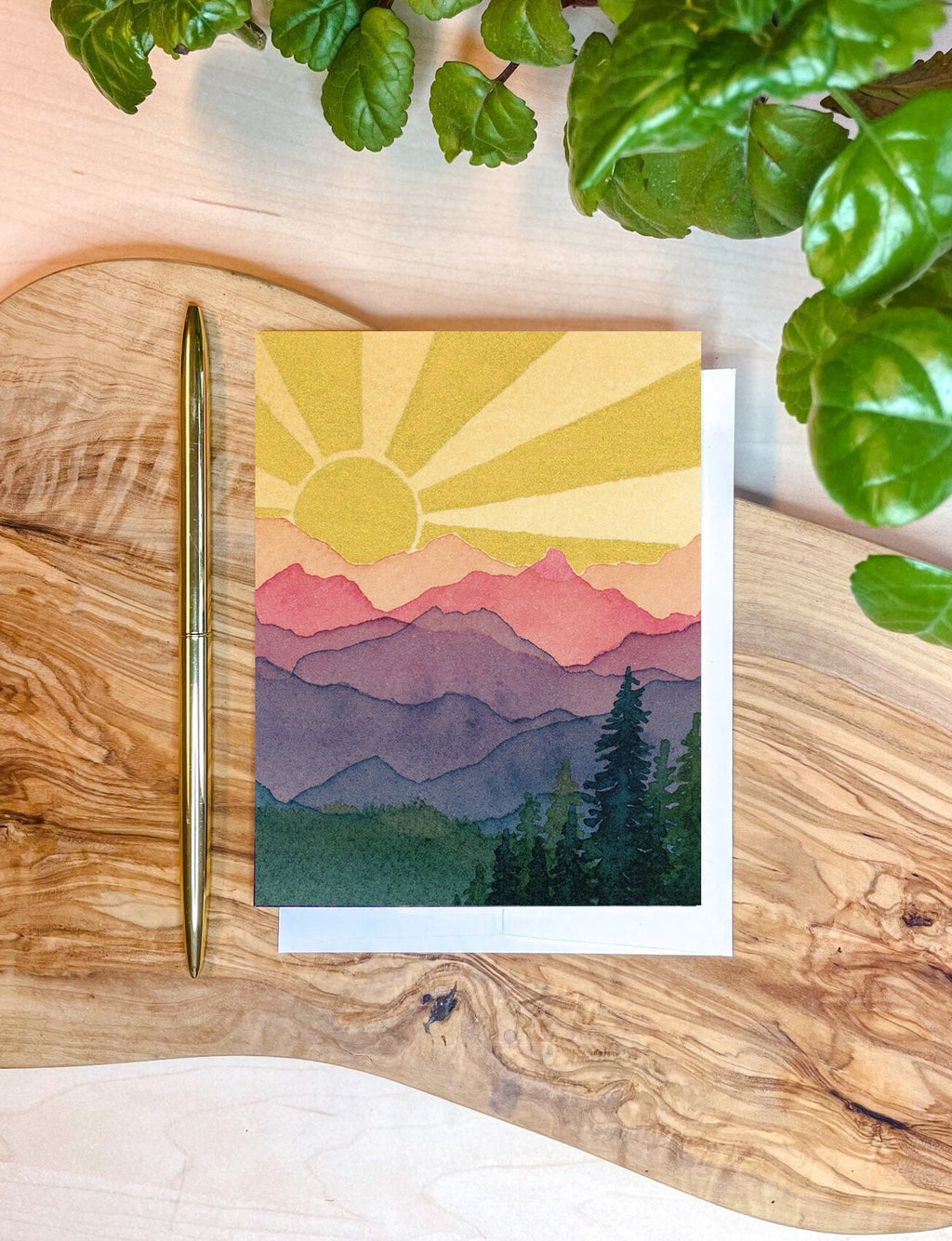 The front of a greeting card with painted mountain landscape and sun with sun rays in the sky. On top of a plain white envelope on a wooden writing surface.