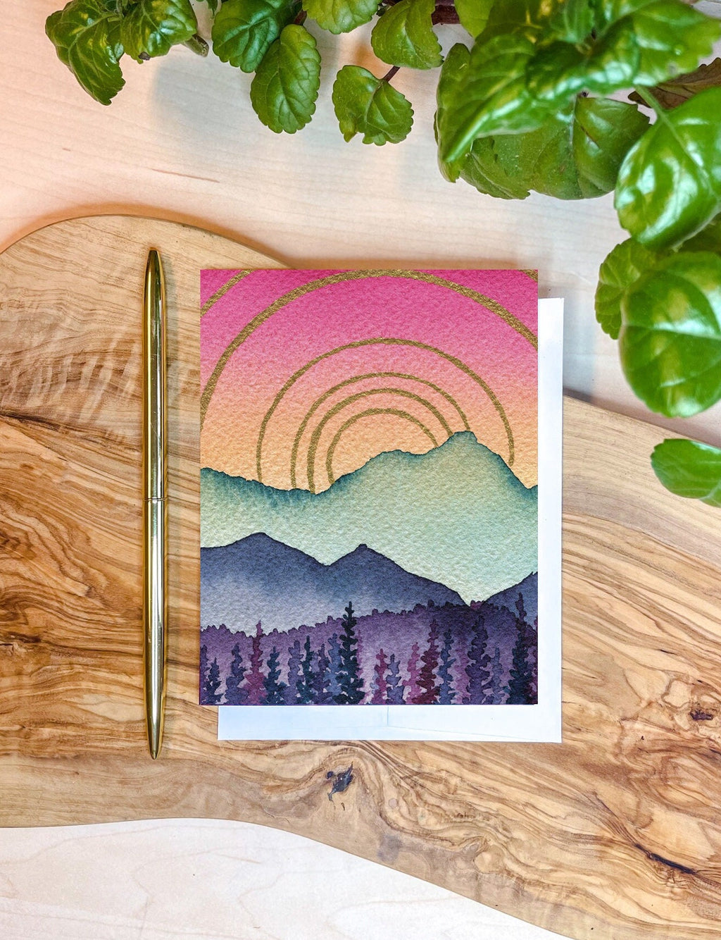 A greeting card with colorful mountain landscape art - a pink and yellow sky with golden rainbows, with mountain layers in turquoise blue and purple. With a white envelope, gold pen, and wooden writing surface.
