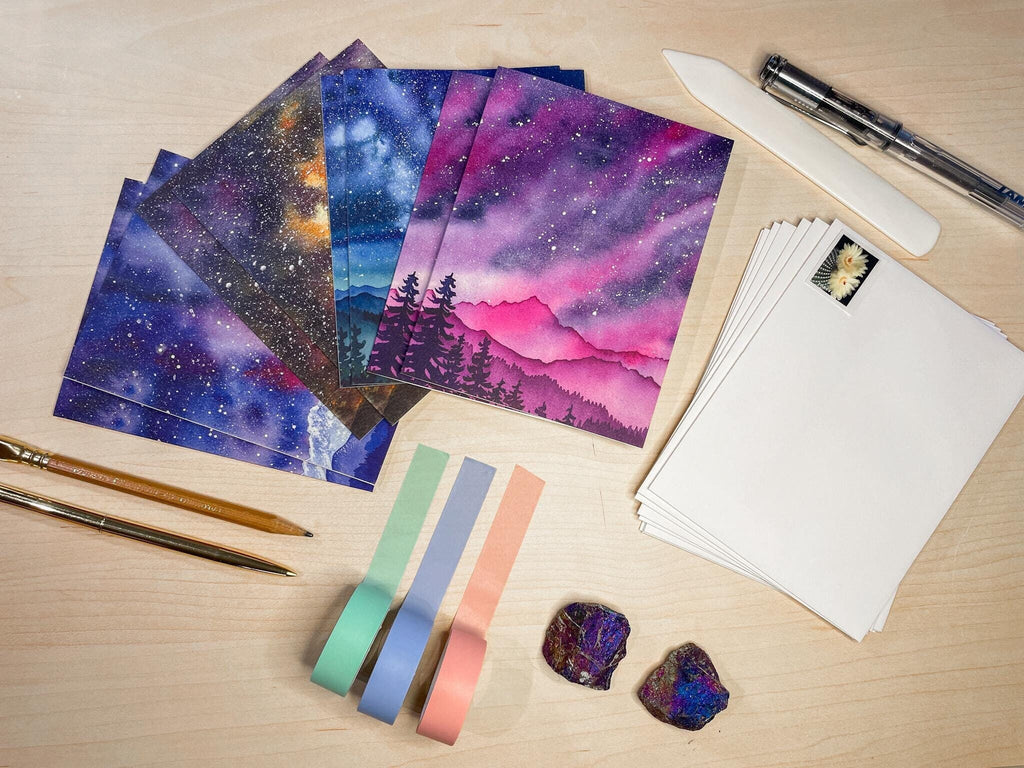 A set of 8 folded greeting card featuring colorful watercolor landscapes with starry skies is spread out on a desk arranged with stationery items.